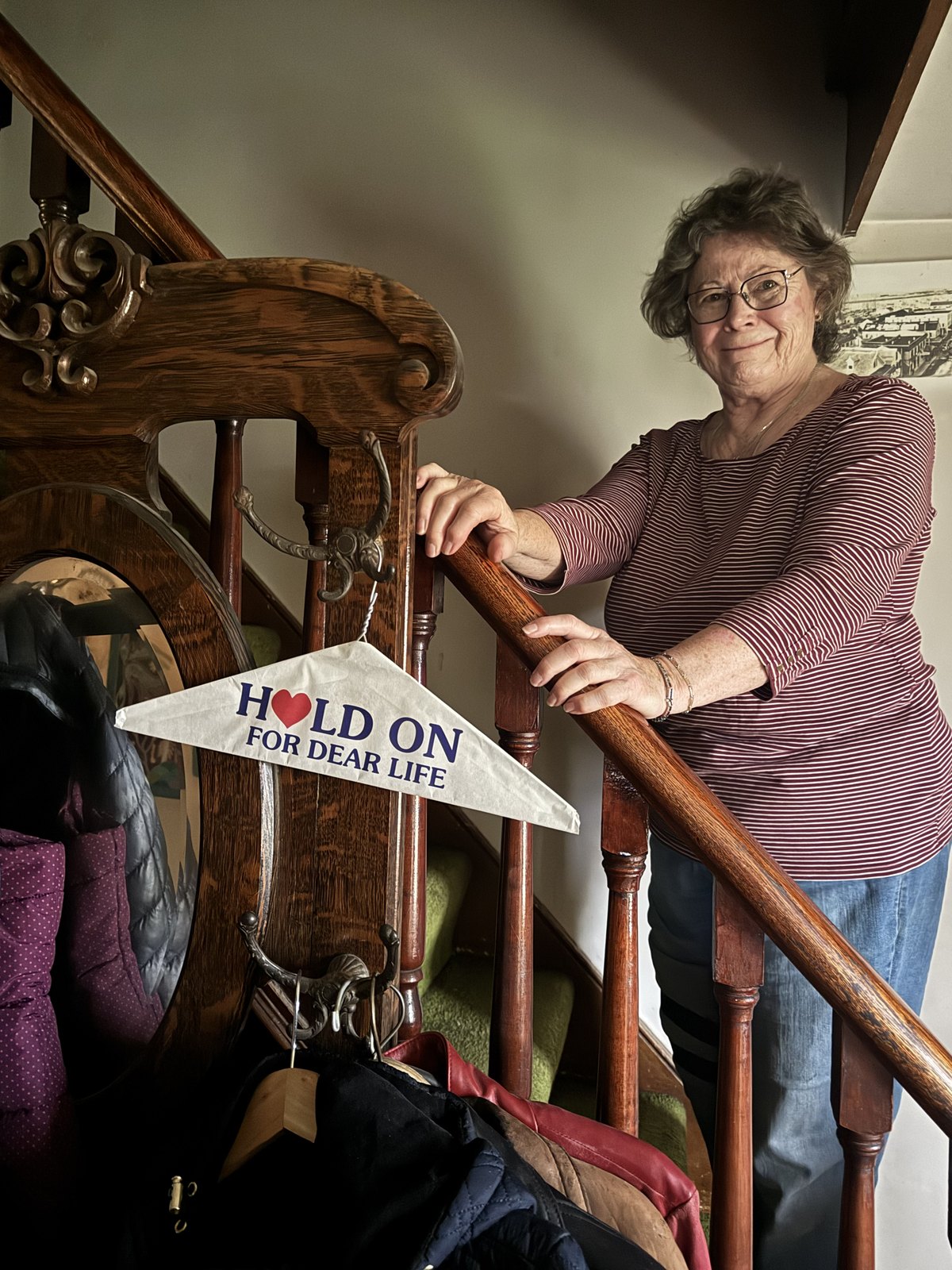 Catherine, Michael's mother, on the staircase at 391 Ontario, holding the banister beside a pennant that reads HOLD ON FOR DEAR LIFE