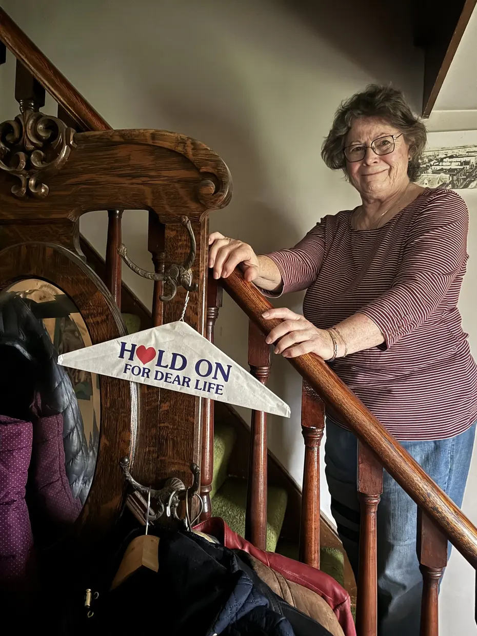 Catherine, Michael's mother, on the staircase at 391 Ontario, holding the banister beside a pennant that reads HOLD ON FOR DEAR LIFE