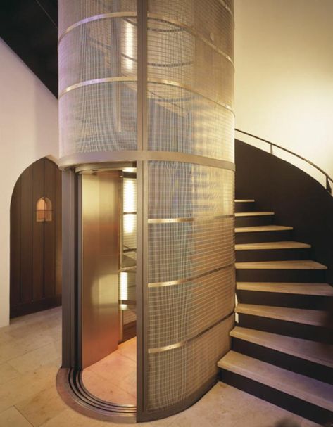A glass-and-mesh residential elevator beside a curved staircase